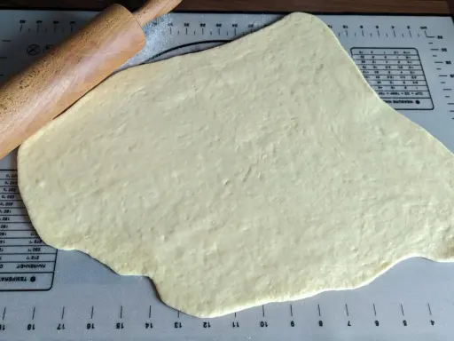 Pâte à beignet légère et croustillante étalée sur un tapis de pâtisserie avec un rouleau à pâtisserie en bois.