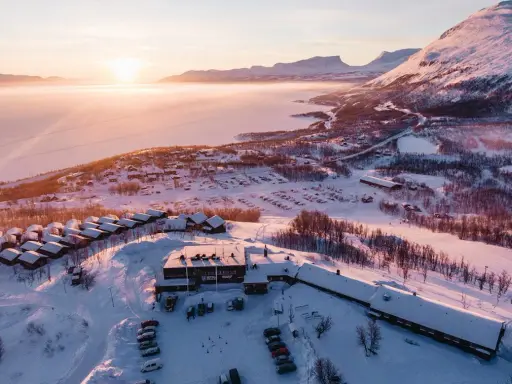 Vinterlandskap vid Kiruna med snötäckta fjäll, en frusen sjö och små stugor. Solen går ner och färgar himlen rosa.