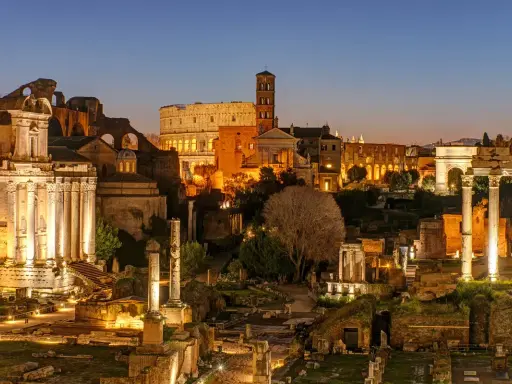 Antike Ruinen des Forum Romanum in Rom bei Nacht. Der Kolosseum und ein Triumphbogen sind beleuchtet.