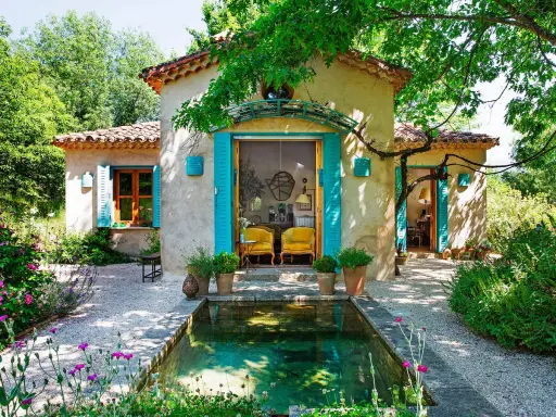 Encantadoras fachadas de casas rústicas con contraventanas turquesas y un estanque rodeado de flores.