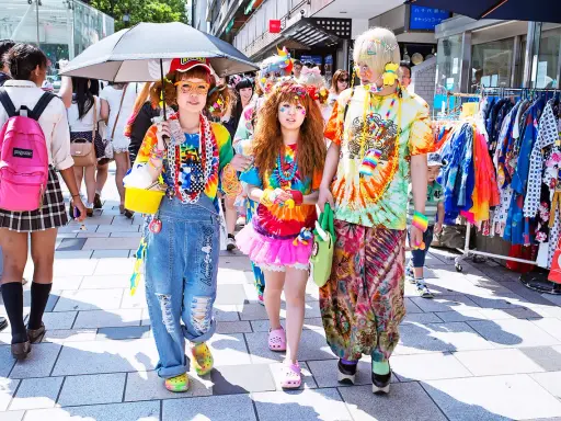 Kolorowy tłum w stylu harajuku, z tęczowymi ubraniami, dżinsowymi ogrodniczkami i jaskrawymi dodatkami, spaceruje ulicą.