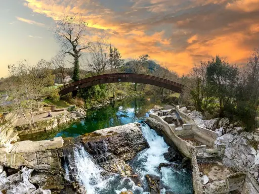 Un puente arqueado cruza un río verde, con cascadas y ruinas de piedra. Un paisaje idílico que invita a descubrir pueblos bonitos cerca de Puente Viesgo.