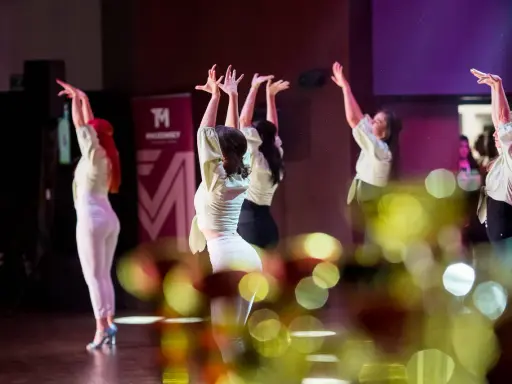 Tancerki w jasnych bluzkach i białych spodniach wykonują zmysłowy taniec. Ich dłonie unoszą się w powietrzu, tworząc dynamiczną choreografię. To piękny pokaz bachata solo.