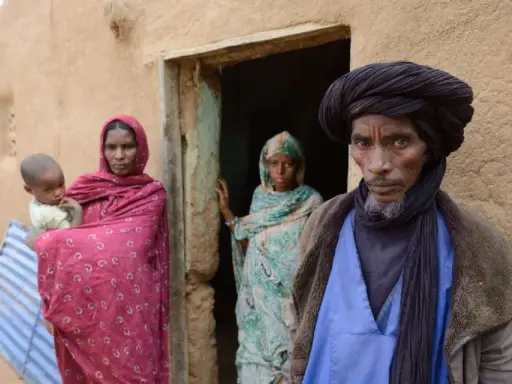 Ein Tuareg-Mann mit schwarzem Turban und ein Kind mit seiner Mutter vor einem Lehmhaus in der Sahara.