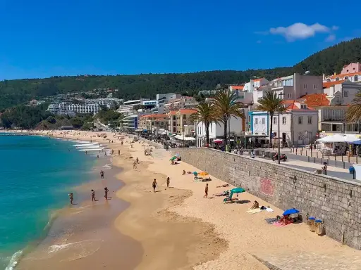 Sonne, Strand und Meer in Sesimbra, Portugal. Menschen genießen den Tag am goldenen Sandstrand, während im Hintergrund die Stadt und grüne Hügel aufragen.