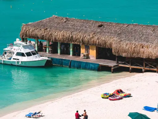 A boat docks at a thatched-roof beach bar on a sunny day. This scene embodies the welcoming spirit of Aruba, a destination known for being gay friendly.