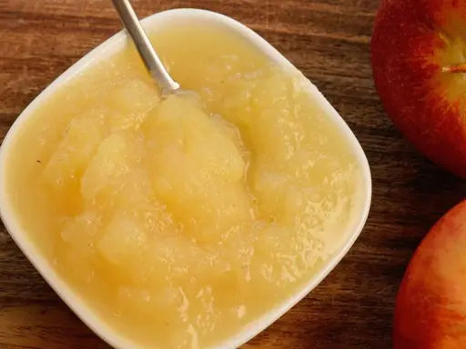 Frisches Apfelmus mit Schale, zubereitet im Thermomix, in einer weißen Schale auf Holzuntergrund. Daneben liegen zwei rote Äpfel.
