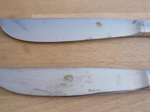 Zwei Messer mit deutlichen Rostflecken auf dem Besteck liegen auf einem Holztisch.