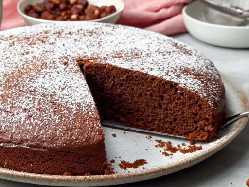 Ein saftiger Nutella Kuchen, bestäubt mit Puderzucker, wartet darauf, angeschnitten zu werden. Daneben eine Schüssel mit Haselnüssen.