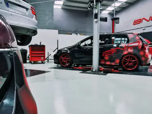 Taller con coches modificados. Uno negro con diseño de hexágonos rojos y llantas rojas, otro negro y rojo con alerón. Se pregunta cuánto cuesta reprogramar un coche.