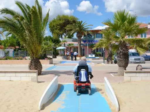 Accesso facilitato per tutti nei villaggi in Abruzzo sul mare. Un uomo in carrozzina elettrica percorre una passerella blu sulla sabbia, tra palme rigogliose e un edificio con la scritta "Welcome".