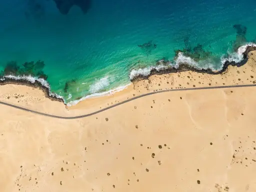 Luftaufnahme eines einsamen **Surferstrands** auf **Fuerteventura**. Türkisblaues Meer trifft auf goldgelben Sand und eine felsige Küste. Eine Straße schlängelt sich entlang der Küste.