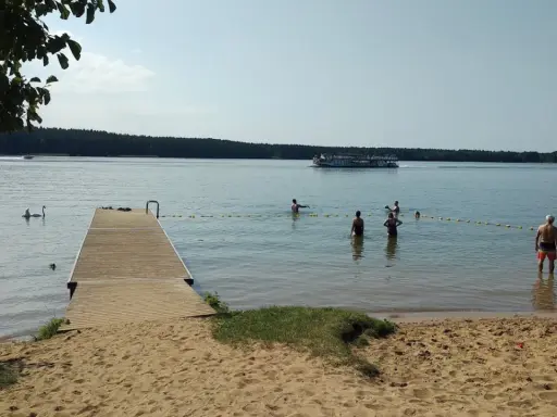 Słoneczna augustów plaża z pomostem, łabędziami i ludźmi kąpiącymi się w jeziorze. Na wodzie pływa statek wycieczkowy.