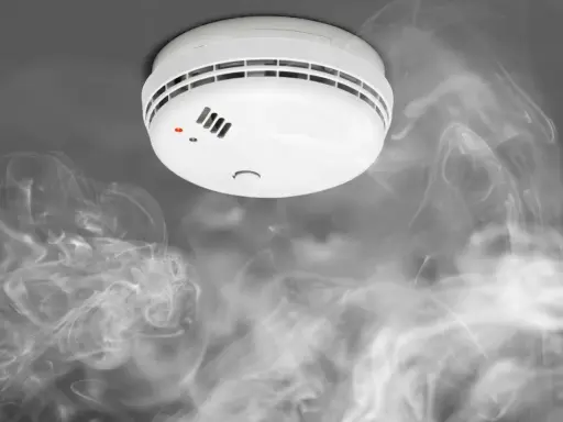 Biały czujnik dymu na suficie, otoczony kłębami dymu. Widać czerwoną diodę sygnalizującą alarm.