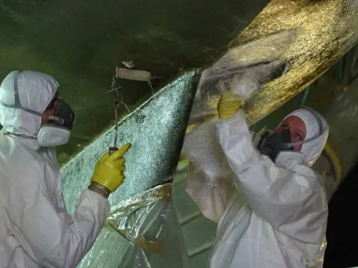 Zwei Personen in Schutzkleidung reparieren den Rumpf eines Bootes. Sie tragen Glasfasermatten auf, um Osmose-Schäden zu beheben.