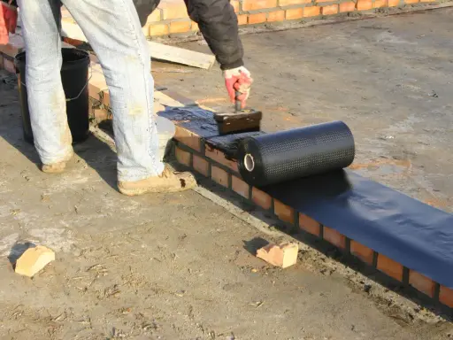 Pracownik nakłada masę bitumiczną na ceglaną ścianę, przygotowując ją do położenia hydroizolacji z rolki.
