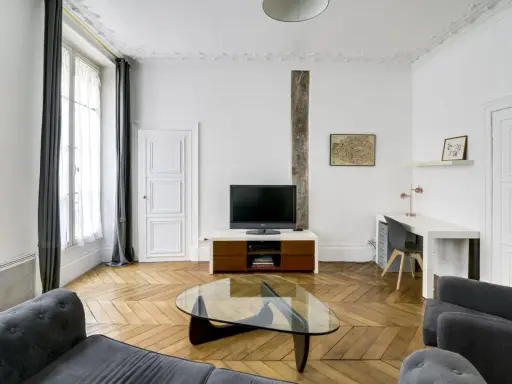 Salon moderne avec canapé gris, table basse en verre, bureau et mur blanc orné d'une carte de Paris.