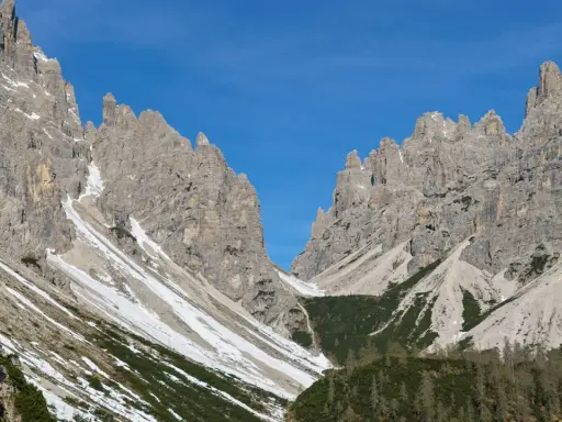 Vette rocciose imponenti sotto un cielo azzurro. Un sentiero scende verso il rifugio Giaf, tra pini e chiazze di neve.