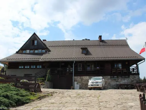 Drewniana bacówka Biała Woda z białym SUV-em na kamienistym podjeździe. Biało-czerwona flaga powiewa na maszcie.