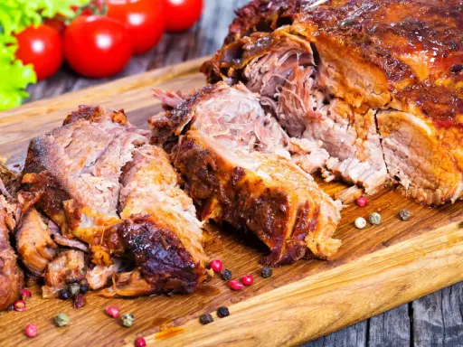 Une succulente épaule d'agneau rôtie, effilochée et servie sur une planche en bois, avec des tomates et de la laitue en arrière-plan.