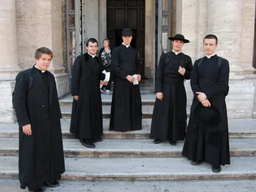 Cinque giovani seminaristi in abiti talari neri, alcuni con cappello, posano sulle scale di un edificio storico.