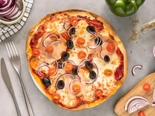 Gotowe ciasto na pizzę z szynką, oliwkami, pomidorkami i czerwoną cebulą. Obok sztućce i miseczka z cebulą.