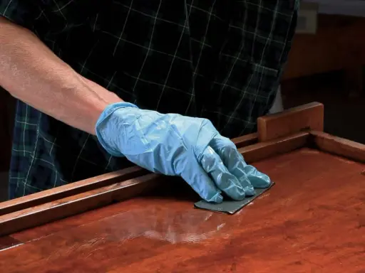 Mano enguantada lijando superficie de madera barnizada. El lijado es un paso clave para aplicar lac.