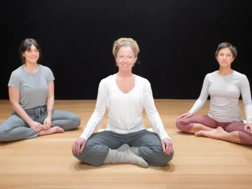 Tre kvinnor sitter i lotusställning på ett trägolv. De deltar i en klass i traumaanpassad yoga.