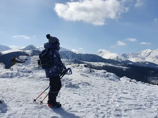 Chłopiec w zimowej kurtce i czapce z pomponem stoi na ośnieżonym szczycie, podziwiając wielki kopieniec gór. W rękach trzyma kijki trekkingowe.