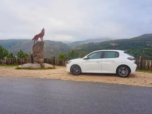 Ein weißer Kleinwagen parkt neben einer Wolfsstatue auf einem Felsen mit Blick auf eine bergige Landschaft. Tolle Carjet Erfahrungen für diese Reise!