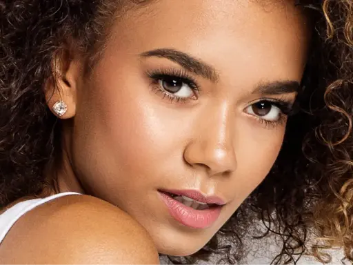 Un gros plan sur le visage d'une jeune femme aux cheveux bouclés, portant une boucle d'oreille étincelante. Ses **rehaucils avis** sont subtils, accentuant son regard.