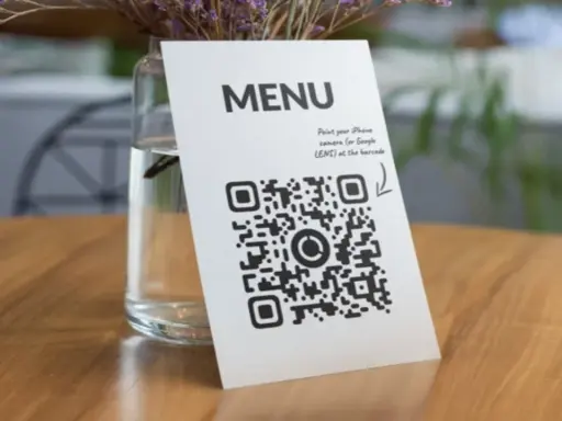 Un código QR de restaurante en una mesa de madera, junto a un jarrón con flores secas. Escanea para ver el menú.
