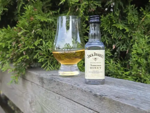 En liten flaska Jack Daniels Honey och ett whiskyglas står på en träbänk, omgivna av grönska.