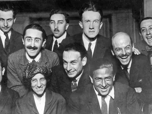 Un gruppo di uomini sorridenti, alcuni con baffi e occhiali, in una fotografia che evoca lo spirito irriverente del dadaismo.