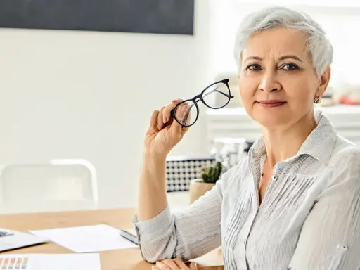 Mujer mayor con gafas, reflexionando sobre los tipos de jubilación. En su escritorio hay un portátil y documentos.