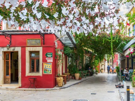 Ein rotes Gebäude mit weißen Tauben-Girlanden über einer Gasse in Chania, Kreta. Tische und Stühle laden zum Verweilen ein.
