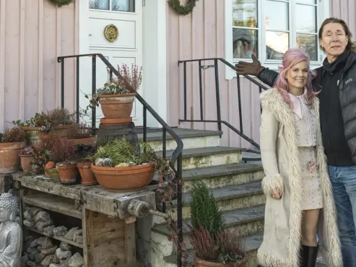 Melinda Jacobs och E-Type poserar glatt framför ett rosa hus med många krukor och en Buddha-staty.