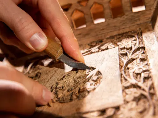 Main d'artisan sculptant un détail floral sur un meuble en bois, étape clé pour savoir comment nettoyer un meuble en marqueterie.
