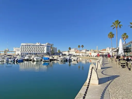 Boote im Hafen von Faro, Portugal, spiegeln sich im ruhigen Wasser. Palmen säumen die Promenade, wo Menschen in Cafés sitzen.