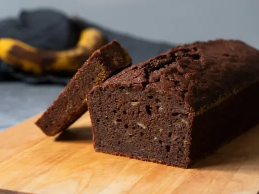 A slice of moist chocolate banana bread is propped against a whole loaf on a wooden cutting board.