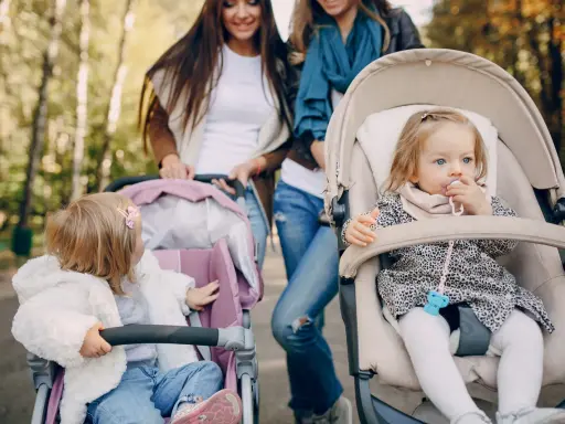 Dwie mamy spacerują z niemowlętami w wózkach. Jedno dziecko ma smoczek, drugie bawi się zabawką. Idealny spacer w parku.