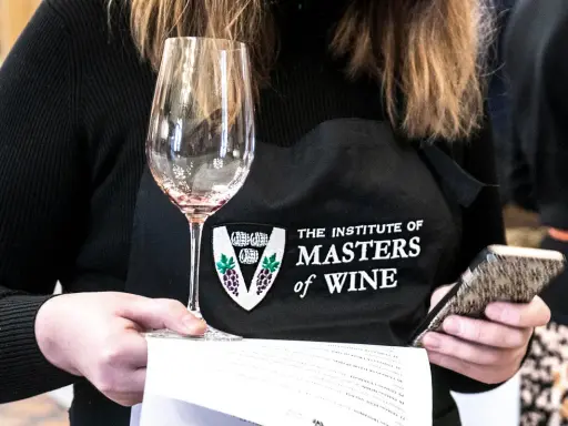 Eine Person mit einer Schürze des "Institute of Masters of Wine" hält ein Weinglas und ein Smartphone.