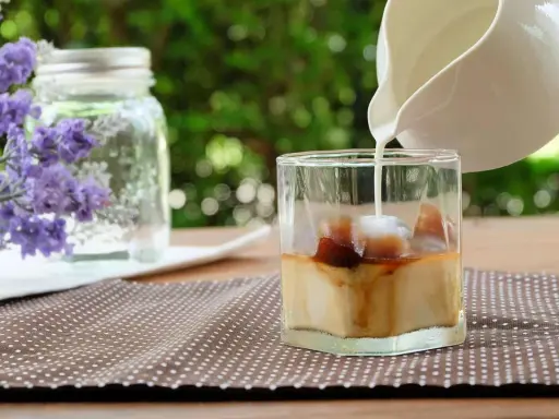 Milch wird in ein Glas mit Eiswürfeln und Kaffee gegossen, um einen erfrischenden Lavendel Latte zuzubereiten.
