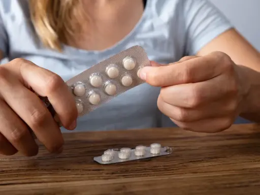Dłonie trzymają opakowanie z tabletkami, które mogą być lekami z serotoniną. Na drewnianym blacie leży drugie, częściowo zużyte opakowanie.