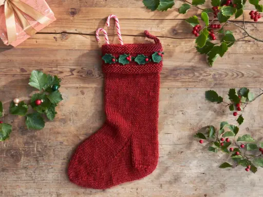 Röd stickad julstrumpa med grön misteldekoration och polkagrisar. En present och julgranspynt ligger på träbordet.