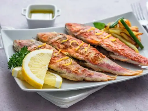 Cuatro salmonetes al horno, dorados y jugosos, servidos con rodajas de limón, perejil y patatas fritas.