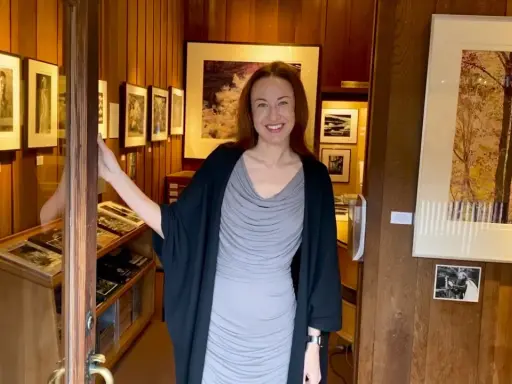 A woman smiles, opening the door to a photography west gallery filled with framed prints and display cases of artwork.