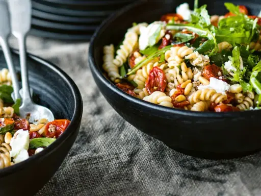 Zwei Schalen mit Nudelsalat mit getrockneten Tomaten, Pinienkernen und Rucola. Ein Löffel liegt in der vorderen Schale.