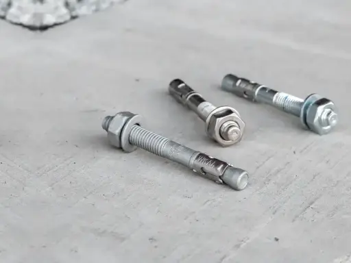 Trzy metalowe kotwy leżą na betonowej powierzchni. Każda kotwa ma gwintowany pręt i nakrętkę.