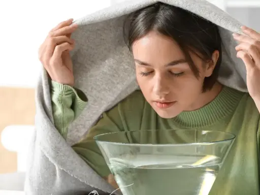 Kobieta z zasłoniętą głową kocem wykonuje inhalację na zatoki nad miską z gorącą wodą.